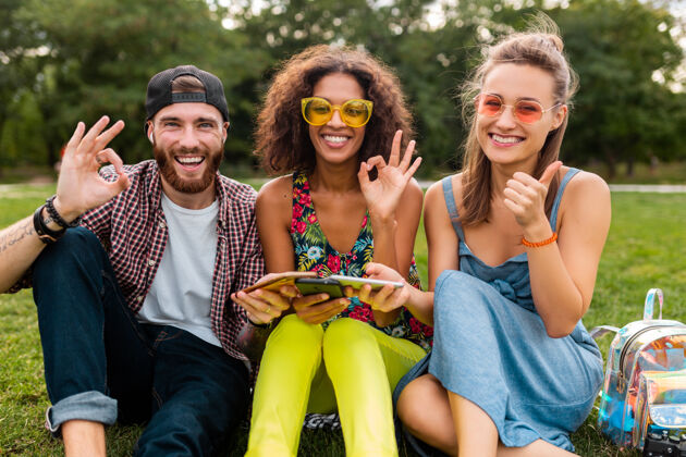 女人快乐年轻的朋友们坐在公园里用智能手机微笑 一起享受快乐多彩的夏日风格 通讯无线连接设备 在相机里看起来很积极时尚人青年