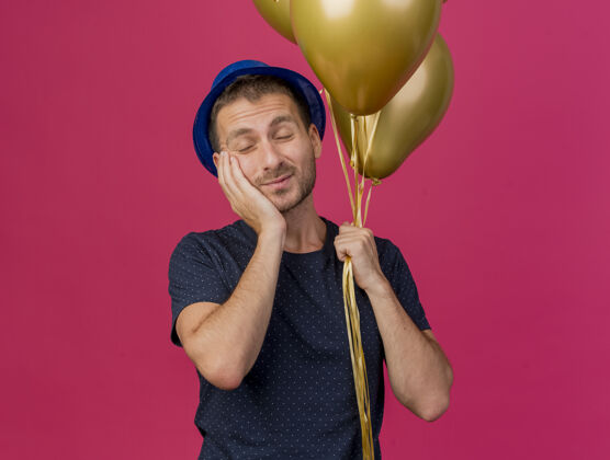 脸戴着蓝色派对帽的白种人帅哥拿着氦气球把手放在脸上 隔离在粉色背景上 留有复制空间粉色复制派对
