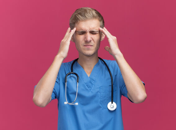 手指年轻的男医生 穿着医生制服 戴着听诊器 把手指放在粉红色墙上孤立的疼痛的太阳穴上太阳穴医生听诊器
