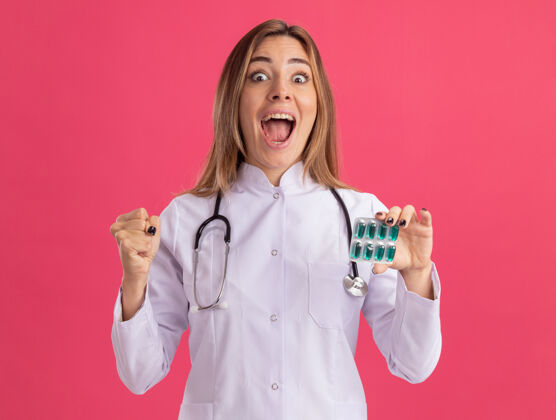 是的兴奋的年轻女医生穿着医用长袍 手持听诊器的药片 在粉红色的墙上显示“是”的手势兴奋长袍手势