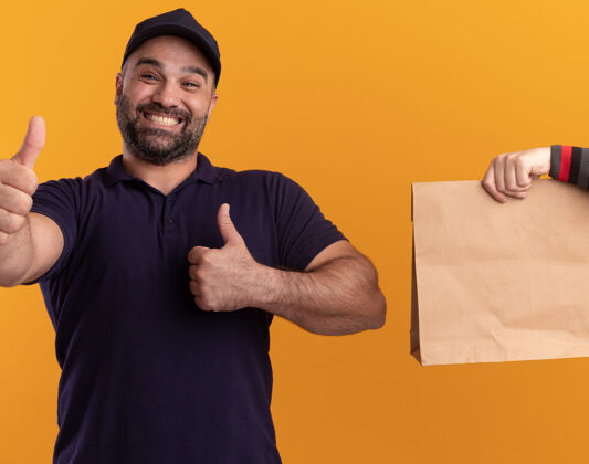顾客微笑着的中年送货员 穿着制服 戴着帽子 向客户赠送纸质食品包装 在黄色的墙上孤立地竖起大拇指拇指帽子制服