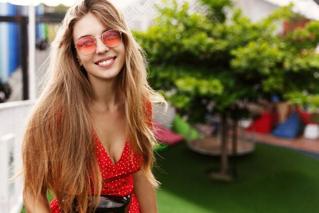 微笑快乐的金发女孩享受热带假期 站在绿色和阳光明媚的公园 微笑着 戴着红色太阳镜女士街道女孩