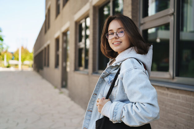 学生现代年轻酷儿女孩的户外写真 戴着眼镜和牛仔夹克的女学生 下课回家 转身对着镜头微笑 等待朋友走在阳光明媚的大街上互联网人大学
