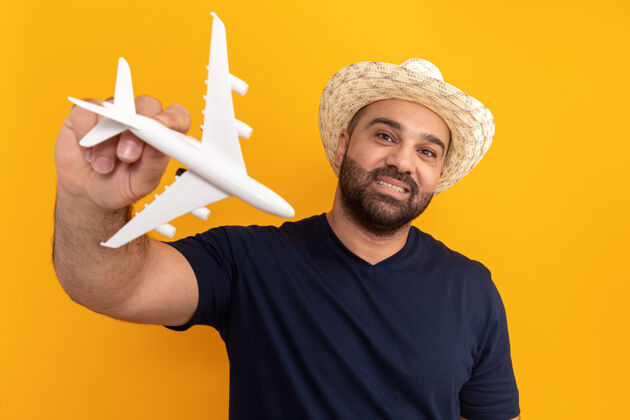 正面留着胡子的男人穿着黑色t恤 戴着夏天的帽子 手里拿着玩具飞机 快乐而积极地微笑着站在橙色的墙上欢呼男人飞机
