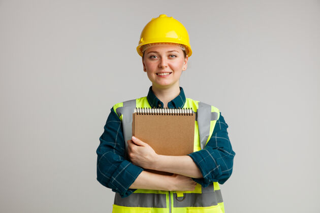 拥抱微笑的年轻女建筑工人戴着安全帽和安全背心拥抱记事本白色头盔佩戴