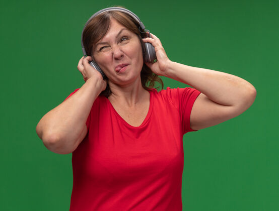 女人中年妇女穿着红色t恤 戴着耳机 伸出舌头 快乐地站在绿色的墙上棍子站舌头