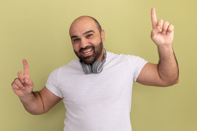 站一个留着胡子的男人 穿着白色t恤 戴着耳机 快乐而积极地微笑 愉快地用食指指着绿色的墙壁目录胡须手指