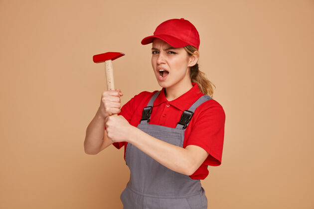 奶油愤怒的年轻女建筑工人穿着制服 手持铁锤帽子穿建筑