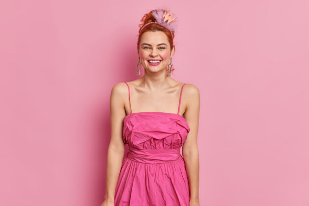 服装时尚的红发年轻女子穿着90年代的风格 穿着粉色的连衣裙 微笑积极 有明亮的化妆姿势 室内准备约会或特殊场合时尚和复古的概念女性姿势魅力