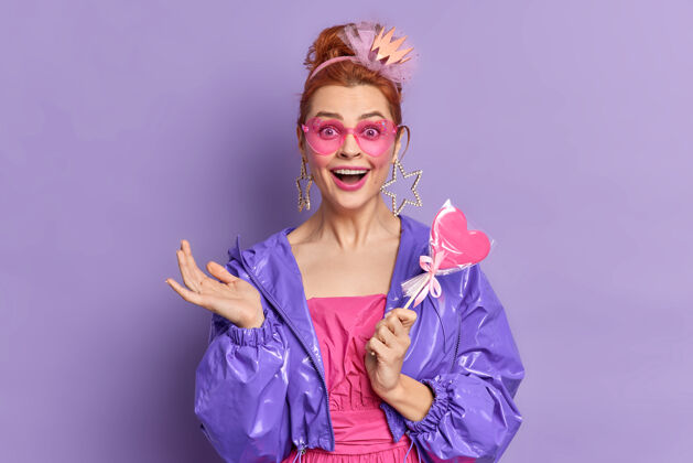 棒棒糖穿着九十年代风格的复古时尚模特有着快乐的表情怀旧在生动的紫色背景下摆着美味的糖果姿势时尚潮流戴着时髦太阳镜的红发女孩紫色夹克积极魅力人类