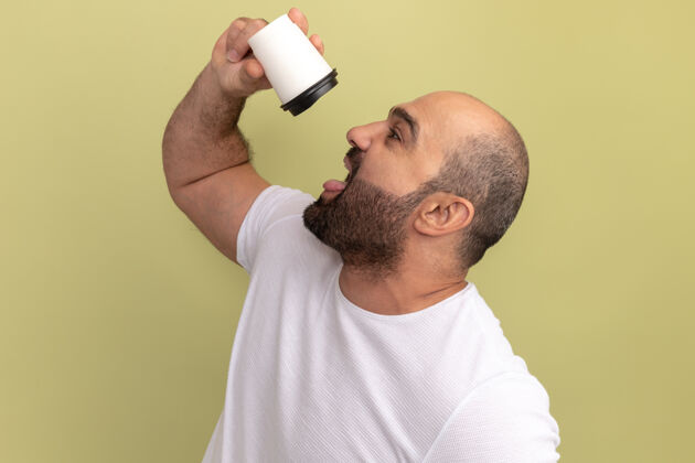 伙计站在绿色的墙上 一个留着胡子的男人穿着白色t恤 手里拿着咖啡杯要喝杯子架子咖啡