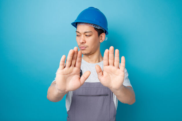 年轻皱眉的年轻建筑工人戴着安全帽和制服 低头做拒绝手势工人皱眉头盔