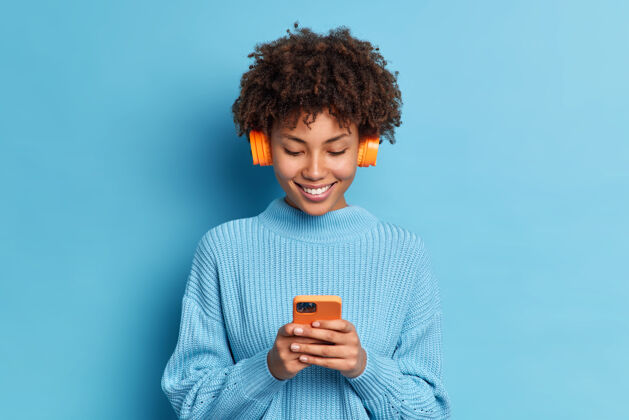 女性身着休闲套头衫的微笑着的非洲头发少年 听着最喜欢的音乐曲目 手里拿着手机下载歌曲到她的播放列表人类青少年手机