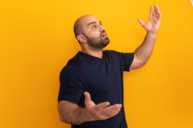 胡子一个留着大胡子的穿海军t恤的男人站在橘色的墙上 困惑地抬头 非常焦虑地举起双臂困惑站起来焦虑