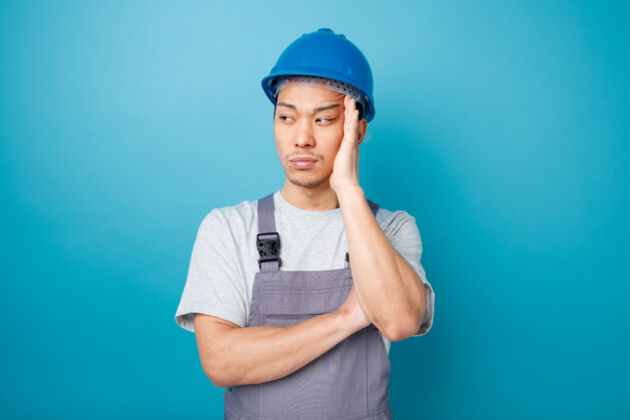 头盔体贴的年轻建筑工人戴着安全帽 穿着制服 手放在头上看着侧面制服穿手