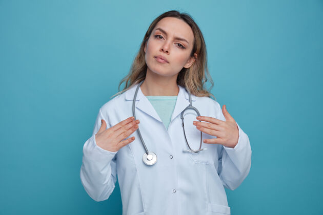 指尖严肃的年轻女医生 穿着医用长袍 脖子上戴着听诊器 用手指着自己手她自己穿
