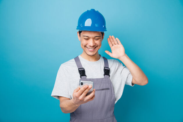 佩戴面带微笑的年轻建筑工人戴着安全帽 穿着制服 手持手机看着挥手建筑蓝色头盔