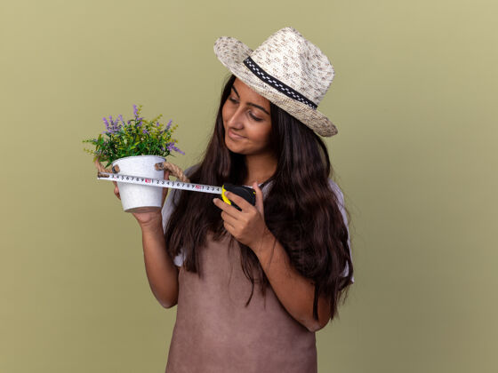 花园穿着围裙 戴着夏帽 拿着卷尺和盆栽植物的年轻园丁女孩站在绿色的墙上 面带微笑地看着植物年轻微笑立场