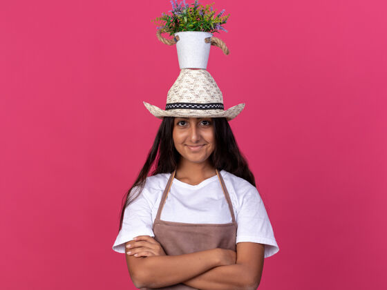 头穿着围裙 头戴夏帽 头戴盆栽的年轻园丁女孩站在粉红色的墙上 脸上带着微笑年轻立场帽子