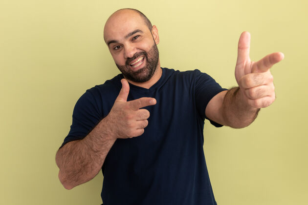 站立一个留着胡子的男人 穿着黑色t恤 脸上挂着快乐的笑容 用食指指着绿色的墙壁表达目录胡须