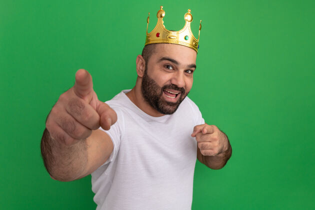 目录戴着金色皇冠的白t恤胡子男人快乐而积极地用食指指着绿色的墙壁指尖佩戴男人