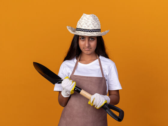 铲子身穿围裙 戴着夏帽 手持铁锹 表情自信的年轻园丁女孩站在橙色的墙上年轻花园表情