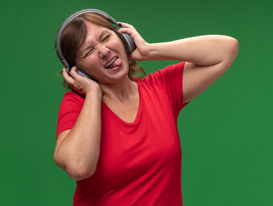 享受快乐的中年妇女 穿着红色t恤 戴着耳机 站在绿色的墙上欣赏她最喜欢的音乐舌头站音乐