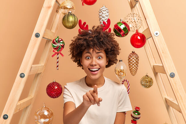 毛衣快乐迷人的卷发民族女子在镜头前直指笑容灿烂准备节日庆典用梯子挂圣诞玩具眼前看到一些神奇的东西指向女性季节