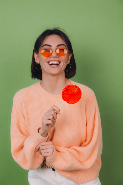 无忧无虑年轻时尚的女士穿着休闲的桃色毛衣 戴着橙色的眼镜 隔离在绿色橄榄色的墙上 带着橙色棒棒糖的正面微笑复制空间酷焦糖青年