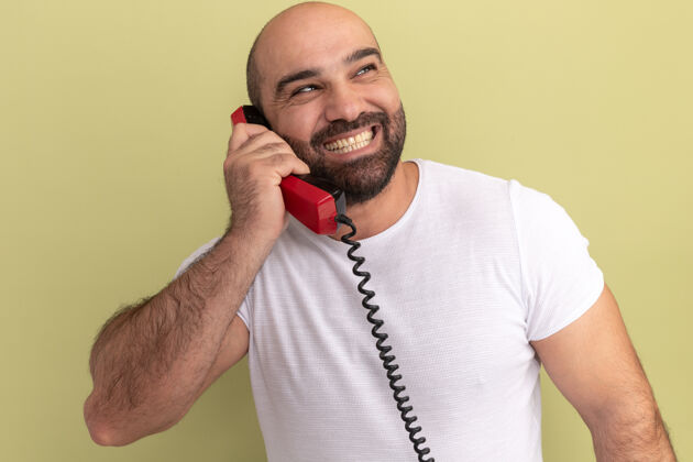 男人一个留着胡子 穿着白色t恤的男人 拿着旧电话 面带微笑 站在绿色的墙上 望向一边靠边站胡子