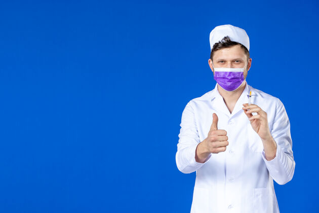 注射身穿医疗服 戴着紫色口罩的男医生正对着蓝色的脸拿着针剂治疗病毒药品