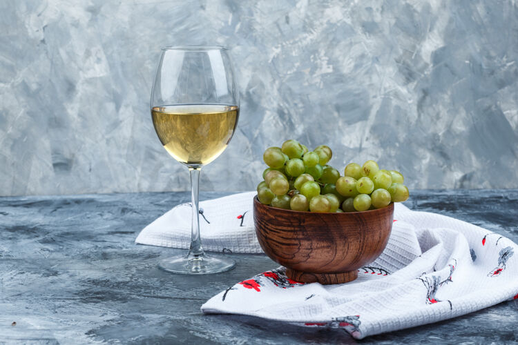 浆果在一个碗里放一杯葡萄酒和绿葡萄 背景是肮脏的石膏和厨房毛巾侧视图水果酒庄大