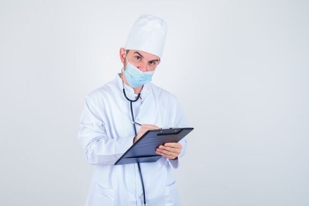 医院年轻的男医生穿着白色制服 戴着口罩 看上去很懂事 正对着剪贴板做笔记智能人士保健人员