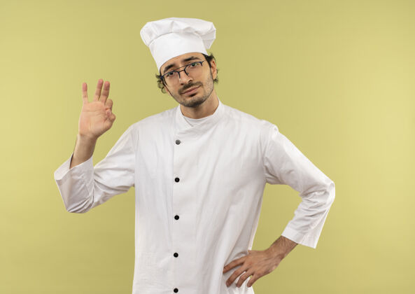 穿年轻的男厨师穿着厨师制服 戴着眼镜 手放在臀部 很高兴制服眼镜臀部