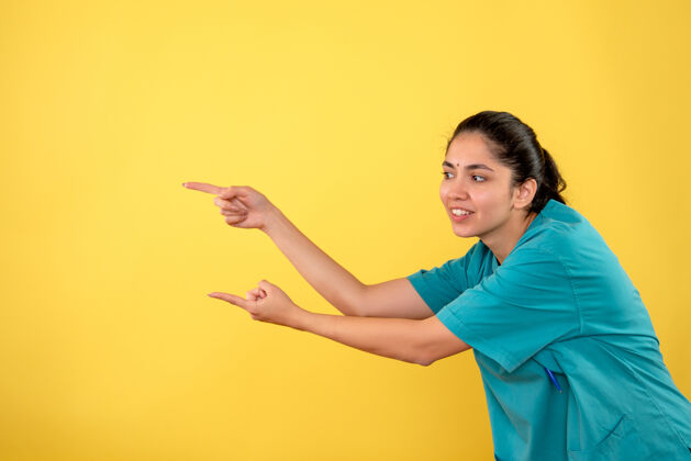 制服正面图黄色背景上穿着制服的快乐年轻女医生用手指向左指模特肖像前面