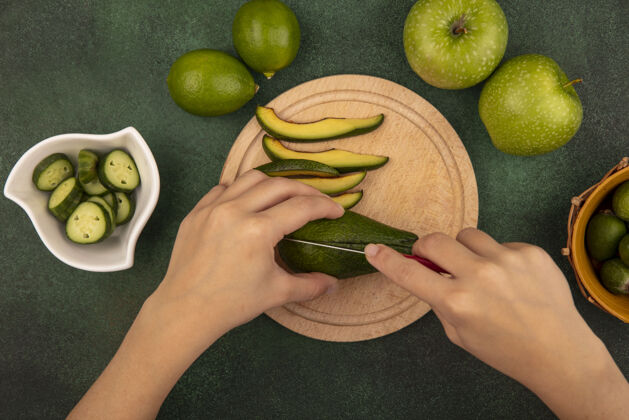 美食俯视图：女性用刀在木制菜板上切鳄梨 切碎的黄瓜片放在碗里 柠檬和青苹果被隔离在绿色的表面上酸橙营养食物