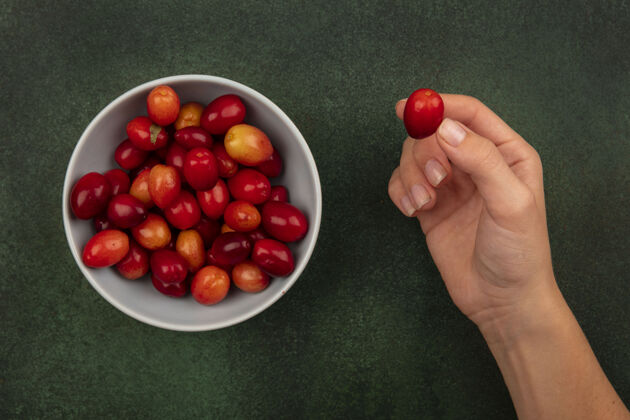 美味俯视图中的女性手拿着一个红樱桃和一碗红樱桃在一个绿色的表面健康碗食物