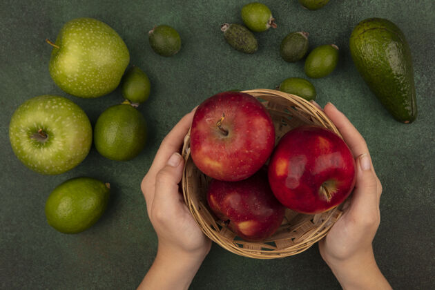 等级俯视图中的女性手拿着一桶新鲜的红苹果和酸橙 绿苹果和绿苹果隔离在一个绿色的表面美味美味饮食