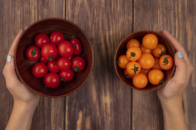 雌性一只手拿着一碗新鲜的樱桃红色西红柿 另一只手拿着一碗橘黄色的樱桃西红柿放在木头表面的女性手的俯视图配料蔬菜农业