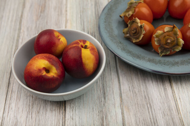 碗一碗美味的桃子和一盘柔软的新鲜柿子放在灰色的木头表面上的俯视图盘子柔软柿子