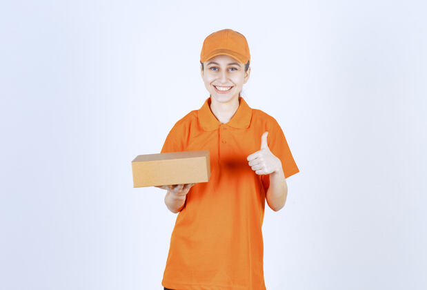 成人穿着黄色制服的女快递员拿着一个纸板箱 手上有肯定的手势强壮订单工人