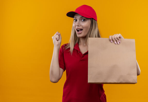 制服快乐的年轻女送货员穿着红色制服 戴着帽子 手里拿着纸袋 摆着“是”的手势展示红色送货