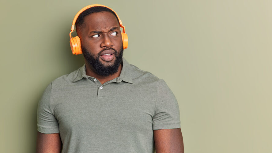 无线迷惑不解的黑胡子男人一边看一边气愤戴着无线耳机一边听音轨姿势穿着休闲t恤隔离在工作室墙上空白的复制空间头男孩旋律