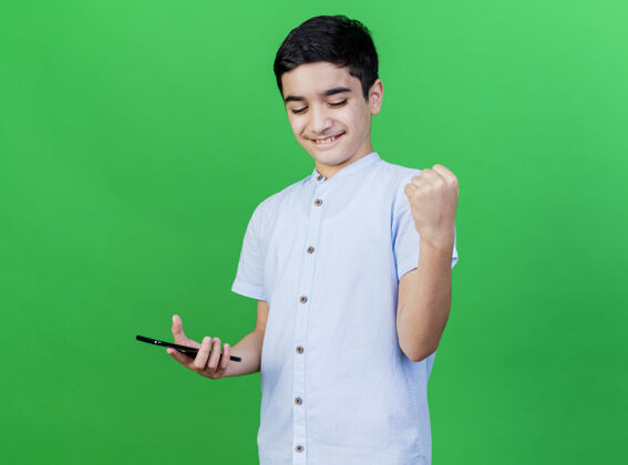 男孩快乐的年轻白人男孩拿着手机做着“是”的手势 隔离在绿色的墙上 留着复印空间快乐电话表达