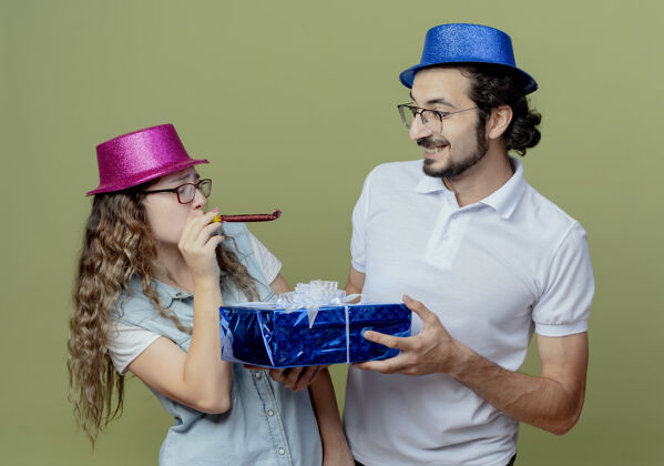 年轻人一对戴着粉色和蓝色帽子的年轻夫妇互相看着吹哨子的女孩 男孩把礼盒送给了被隔离在橄榄绿墙上的女孩给穿上帽子
