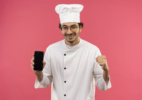 握着面带微笑的年轻男厨师穿着厨师制服 戴着眼镜 拿着电话 在粉红色的墙上显示“是”的手势电话烹饪制服