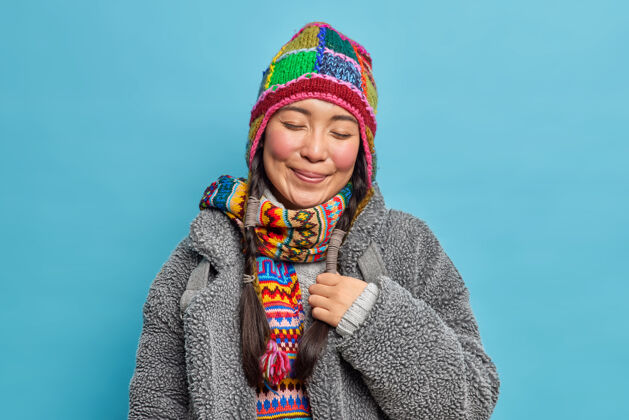 人类满意高兴的亚洲女人闭上眼睛 微笑愉快地戴着针织帽子和围巾寒冷的冬天温暖的外套有两个辫子回忆一些愉快的隔离在蓝色的墙上温暖人季节