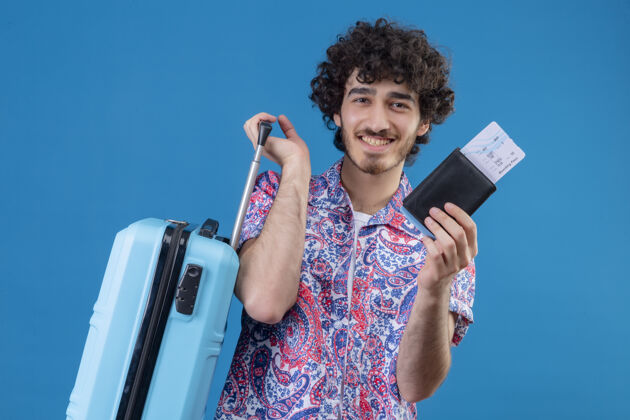 年轻人微笑着的年轻英俊的旅行家 手拿手提箱 机票和钱包 站在孤零零的蓝色墙上抱着微笑手提箱