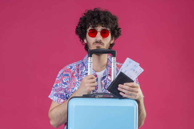 抱着年轻英俊的卷发旅行者戴着太阳镜拿着钱包和机票把手臂放在箱子上孤立的粉红色墙上手提箱胳膊卷发