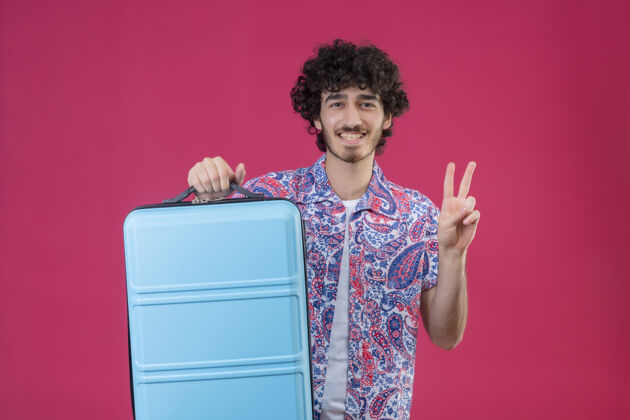英俊微笑着的年轻英俊的卷发旅行者拿着手提箱 在隔离的粉色墙上做着和平的标志 还有复制空间微笑和平卷发